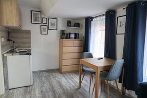 Eine kompakte Küche mit Mikrowelle, Kaffeemaschine und einem kleinen Kühlschrank. Ein Holztisch mit zwei Stühlen und ein blauer Vorhang vor einem Fenster sind ebenfalls vorhanden.