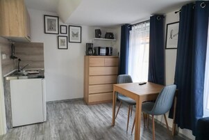 Cocina compacta con microondas, cafetera y refrigerador pequeño. Hay una mesa de comedor de madera con dos sillas y una cortina azul cubriendo una ventana.