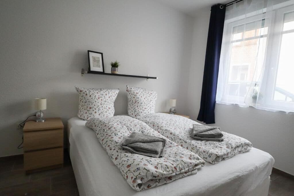 A bedroom with a bed, bedside tables, a shelf with a picture and a plant, and a window with curtains.