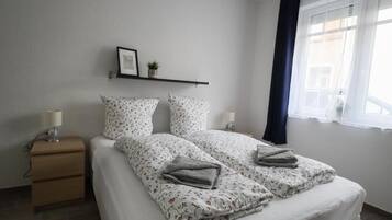 Een slaapkamer met een bed, nachtkastjes, een plank met een foto en een plant, en een raam met gordijnen.