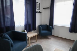 Ein Zimmer mit einer Couch, einem Sessel, einem kleinen Tisch und einem Fenster mit Vorhängen.