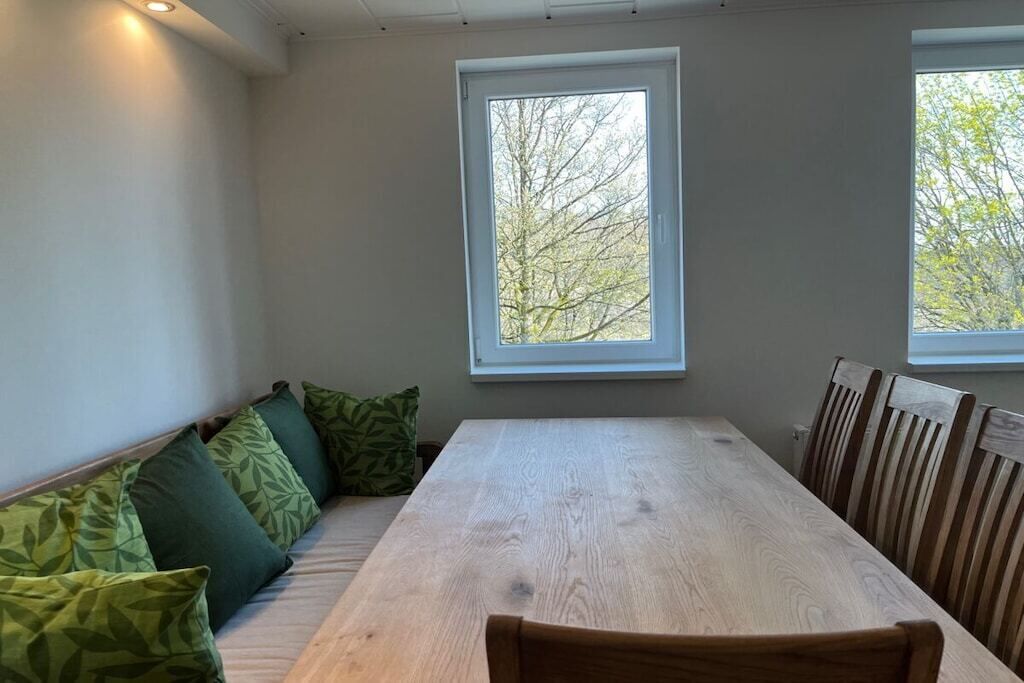 Un espace repas comprenant une longue table en bois et des chaises, un banc avec des coussins et deux fenêtres donnant sur la forêt.