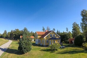 Una casa groga amb teulada vermella, envoltada d'arbres i un jardí amb gespa.
