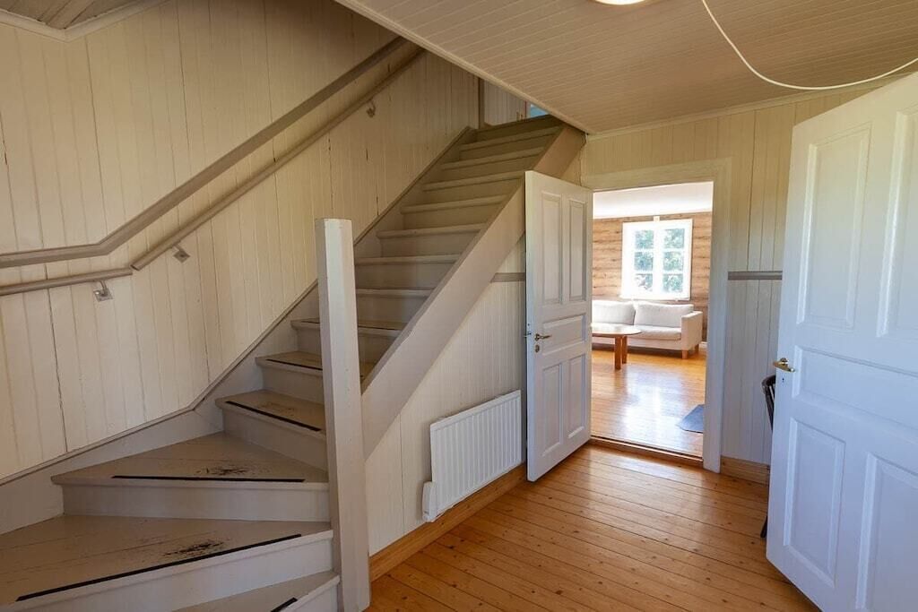 Un escalier mène à une pièce dotée d’une porte blanche, d’un plancher en bois et offrant une vue sur un salon avec un canapé.
