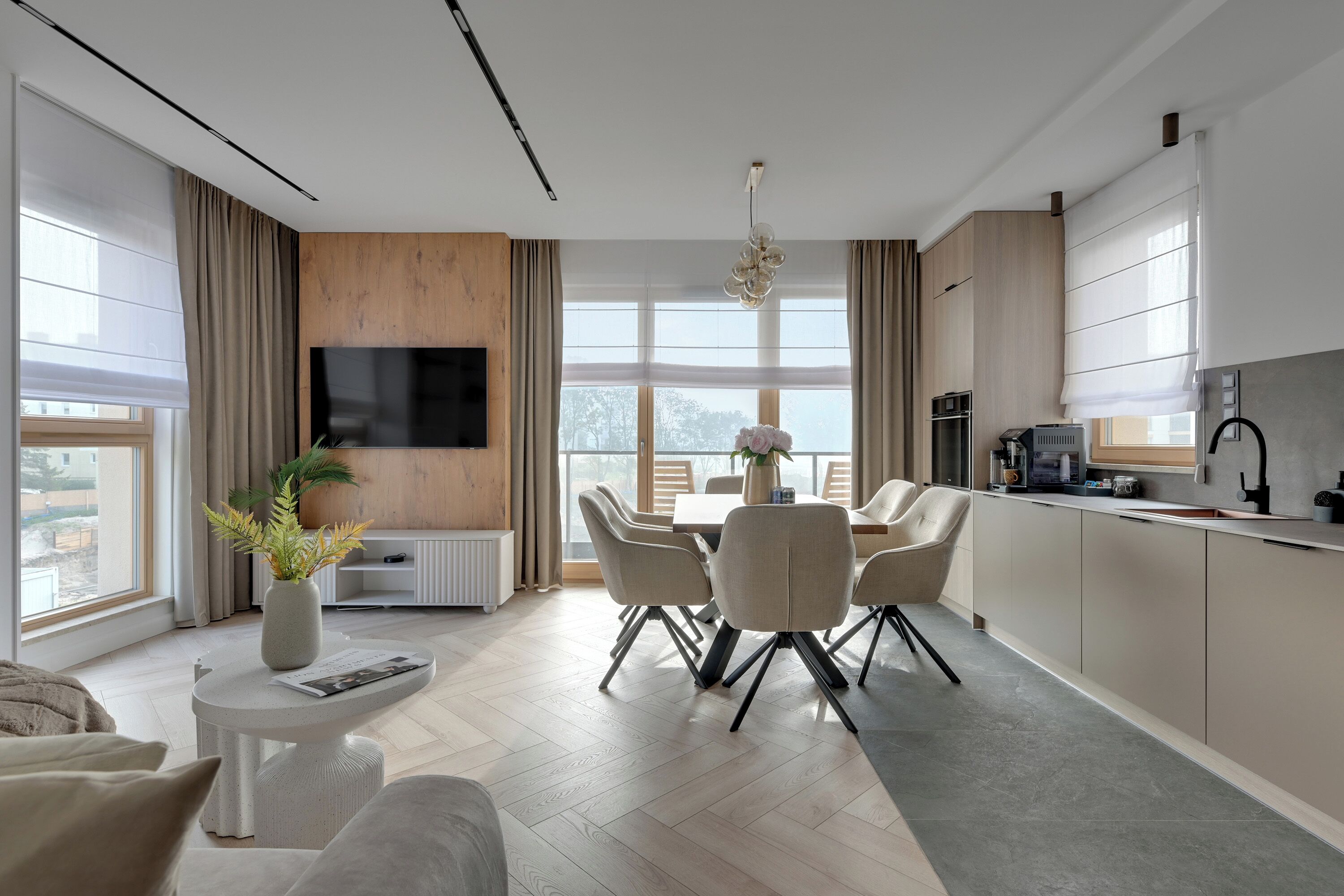 A modern kitchen with a dining area, a flat-screen TV mounted on the wall, and a view of the outdoors.