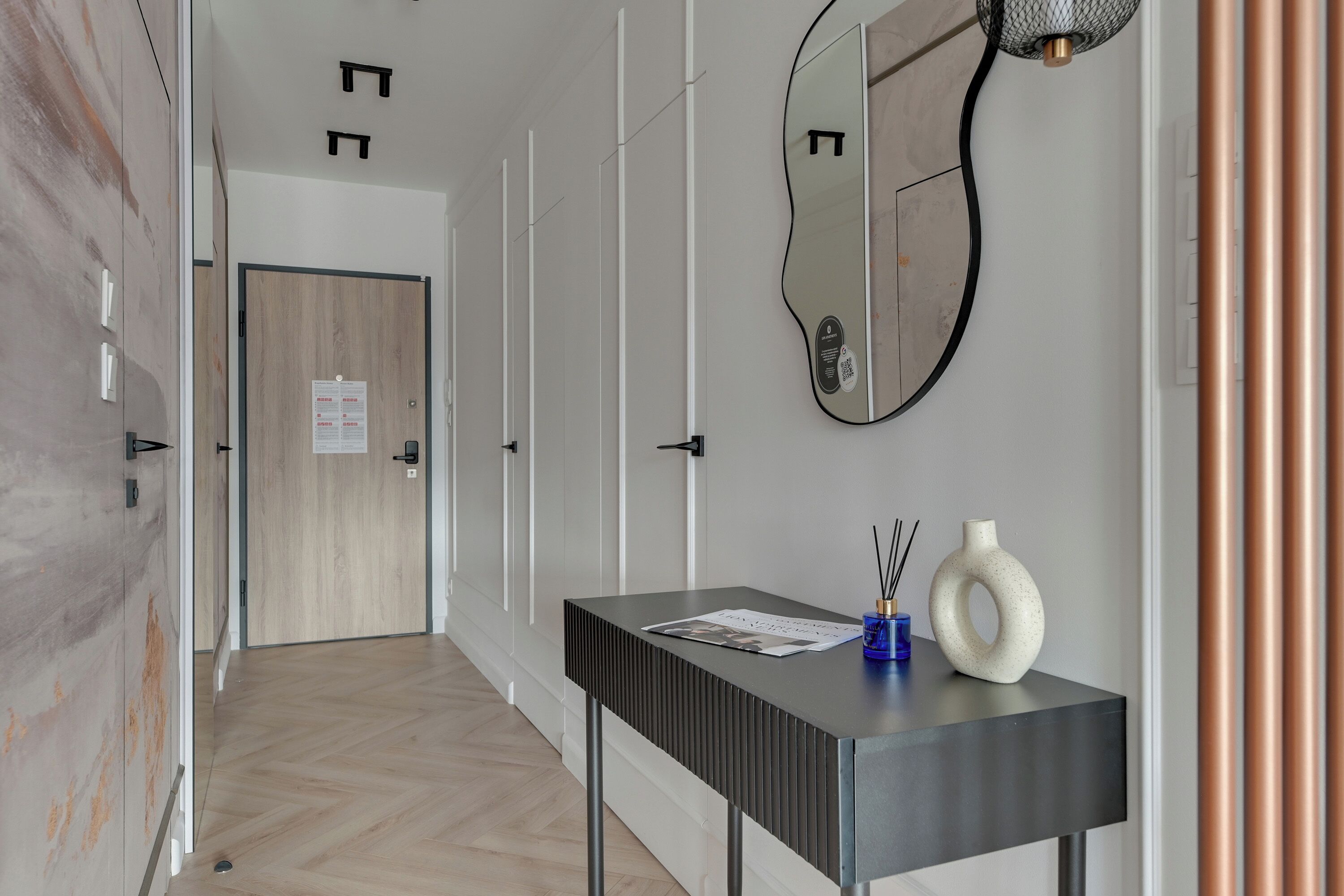 A hallway with a wooden door, a mirror, a black console table with a vase and a candle, and a wall-mounted light fixture.