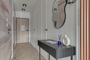A hallway with a wooden door, a mirror, a black console table with a vase and a candle, and a wall-mounted light fixture. - Porto Seaside with Gym and Parking (Gdańsk)