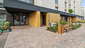 Um edifício moderno com telhado plano, grandes janelas e área de entrada pavimentada, com um banco e um jardim.