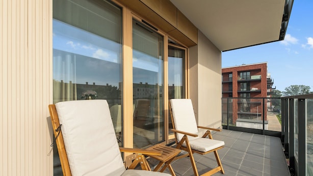 Varanda com duas cadeiras de madeira e uma mesa, janelas amplas e vista para um edifício.