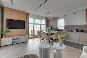 A modern kitchen with a dining area, a flat-screen TV mounted on a wooden wall, and a large window providing natural light. - Porto Seaside with Gym and Parking (Gdańsk)