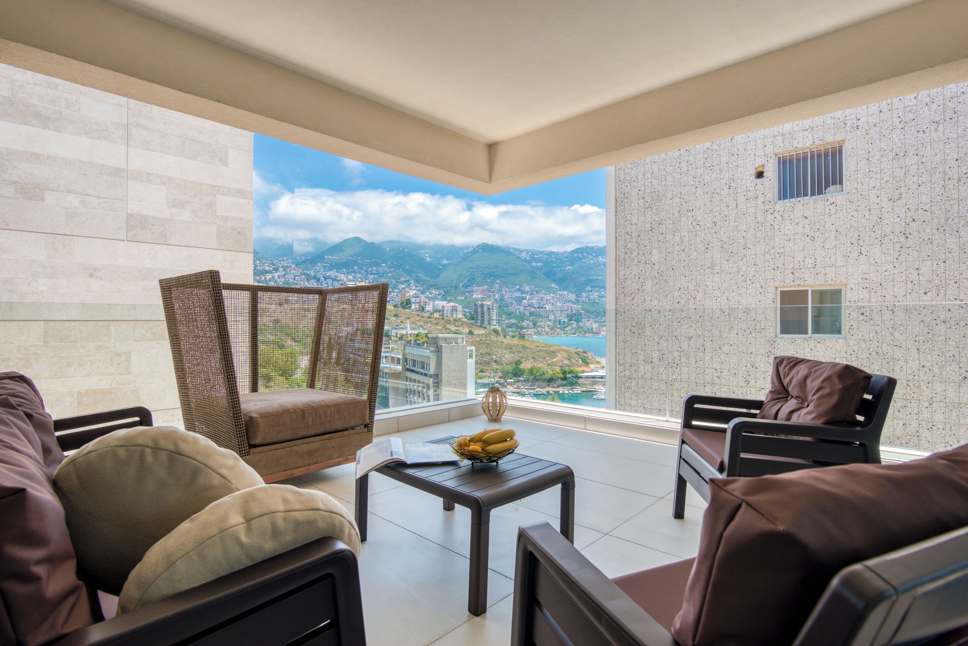 A balcony with a view of a cityscape and mountains, furnished with a sofa, chairs, and a table.