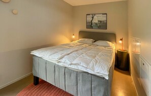A neatly made bed with a grey headboard and white bedding, a small bedside table with a lamp, and a framed picture on the wall. - 2 bedroom beautiful home in Kinnarumma (Kinnarumma)