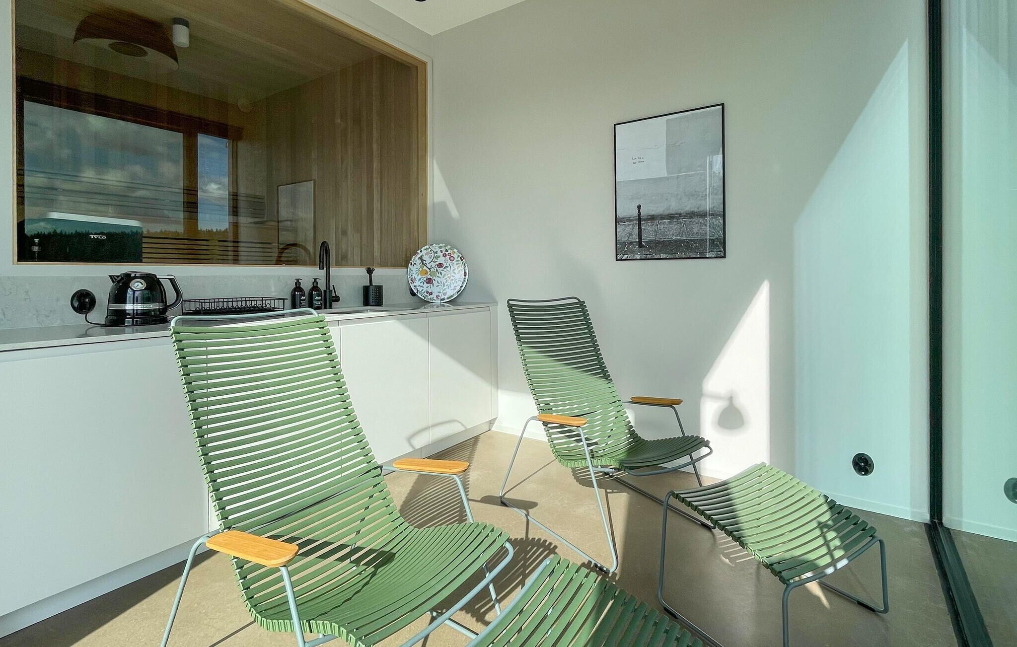 Une cuisine moderne avec une fenêtre, un évier et une bouilloire. On y trouve trois chaises vertes avec des assises en bois.