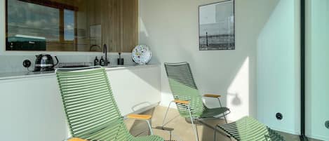 Une cuisine moderne avec une fenêtre, un évier et une bouilloire. On y trouve trois chaises vertes avec des assises en bois.