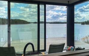 Ein Zimmer mit großen Fenstern mit Blick auf einen ruhigen See, einer Holzterrasse und Sitzgelegenheiten im Freien.