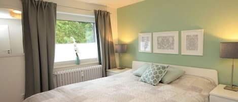 A bedroom with a bed, two bedside tables, a window with curtains, and three framed pictures on the wall.