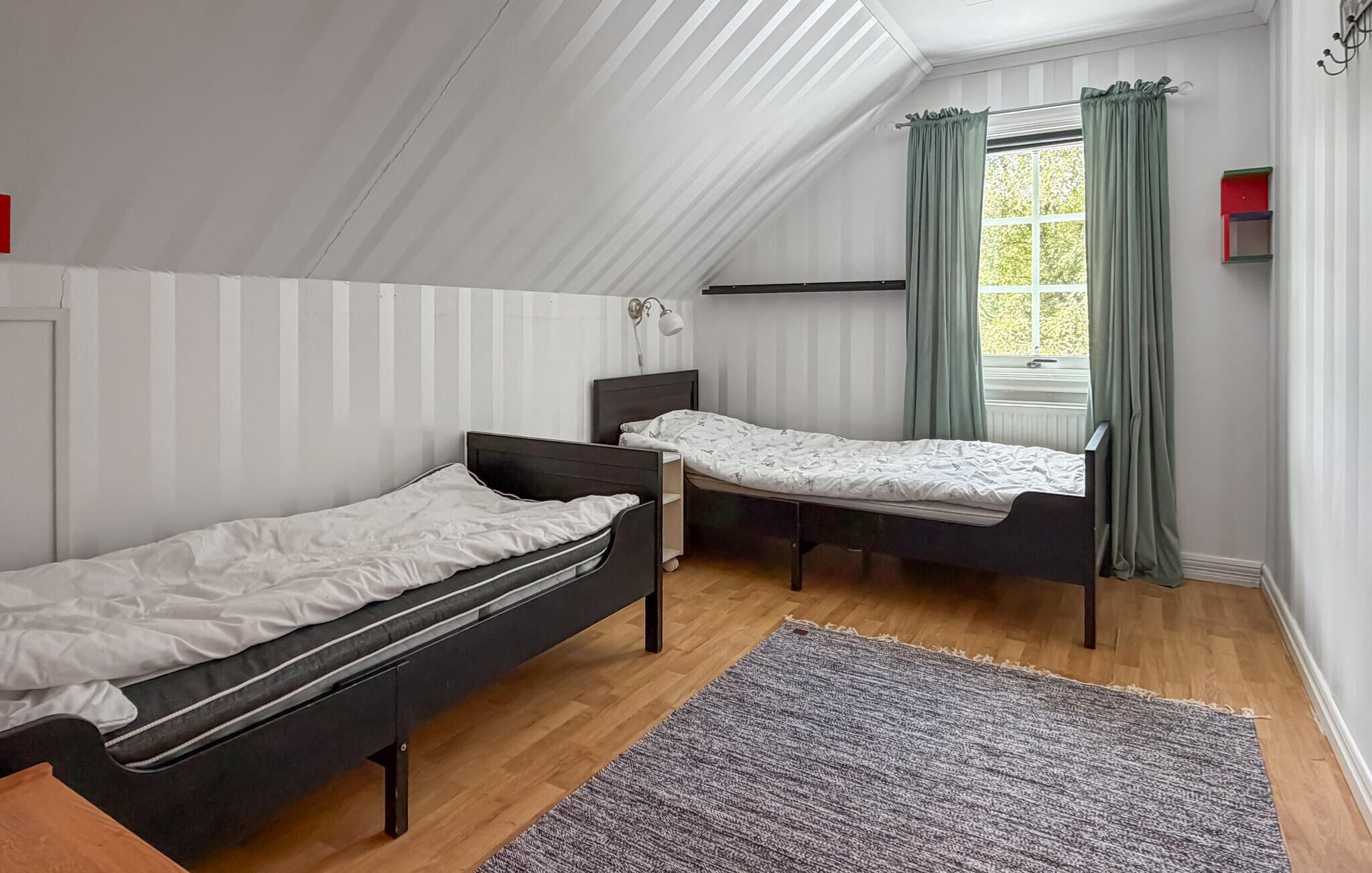 A bedroom with two single beds, a window with curtains, a small bedside table, and a wall-mounted lamp.