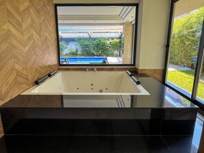 A modern bathroom with a large bathtub, a mirror above it, and a view of a swimming pool and greenery outside. - Qoople Cleopatra Exclusive - Cozy 1-bedroom apartment in Alanya (Alanya)