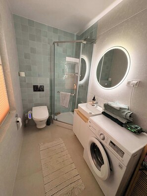 Uma casa de banho moderna com chuveiro em cabine de vidro, sanita, lavatório com espelho e máquina de lavar roupa.