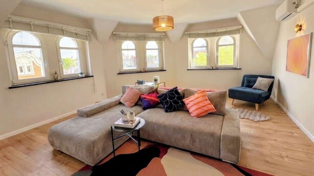 A modern living room with a sectional sofa, a small side table, and a floor lamp. There are large windows with white frames and a view of the outside. - Spacious apartment in central Zielona Gora (Zielona Góra)