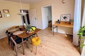 A dining area with a wooden table, chairs, and a yellow cushion. There is a door leading to another room, a shelf with books and a framed picture, and a plant on the right.