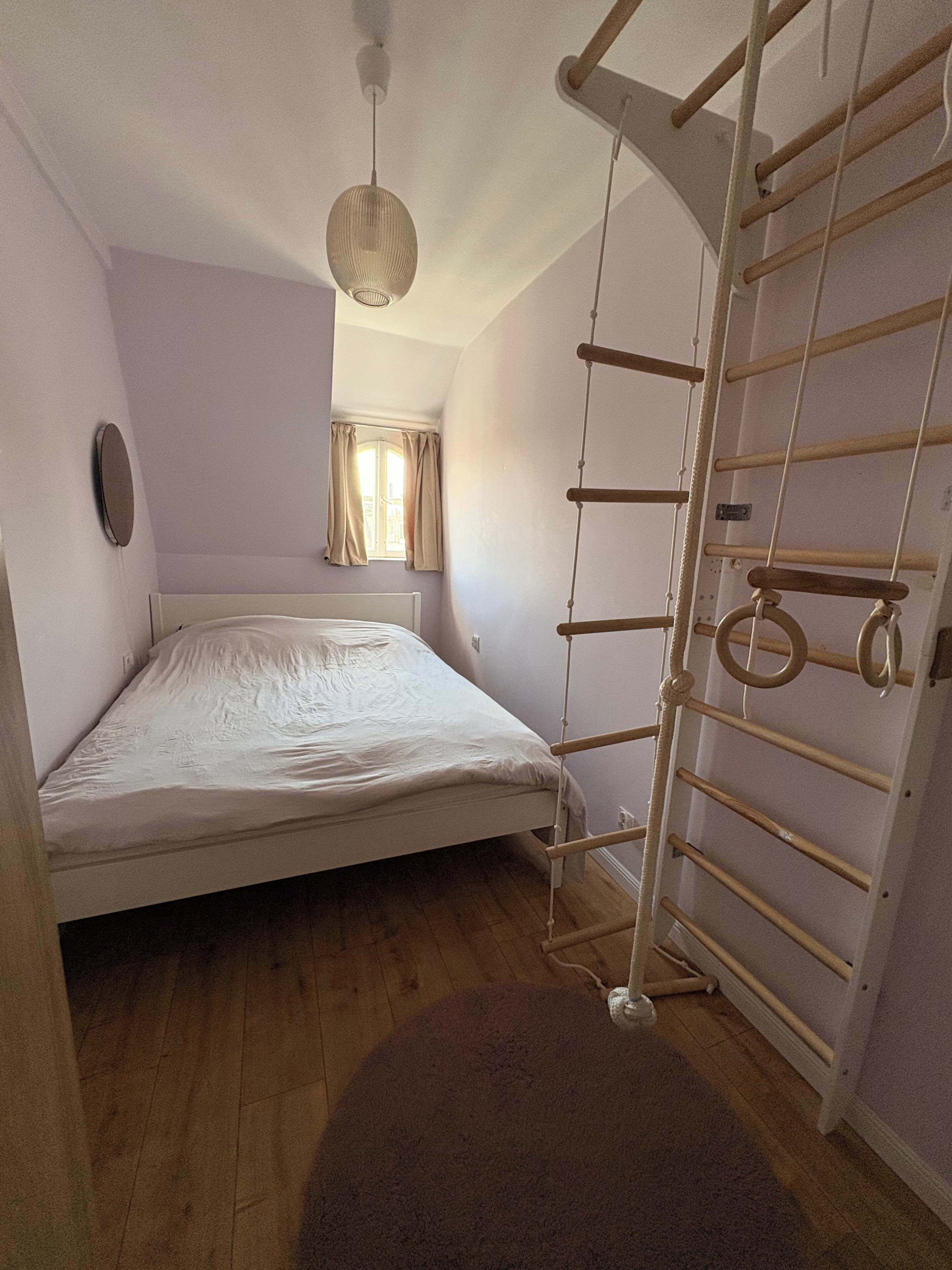 Un lit simple avec une tête de lit, une fenêtre avec des rideaux, un luminaire au plafond et un étagère en métal.