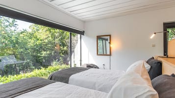 Een slaapkamer met twee bedden, een raam met uitzicht op groen, een spiegel aan de muur en een bedlampje.