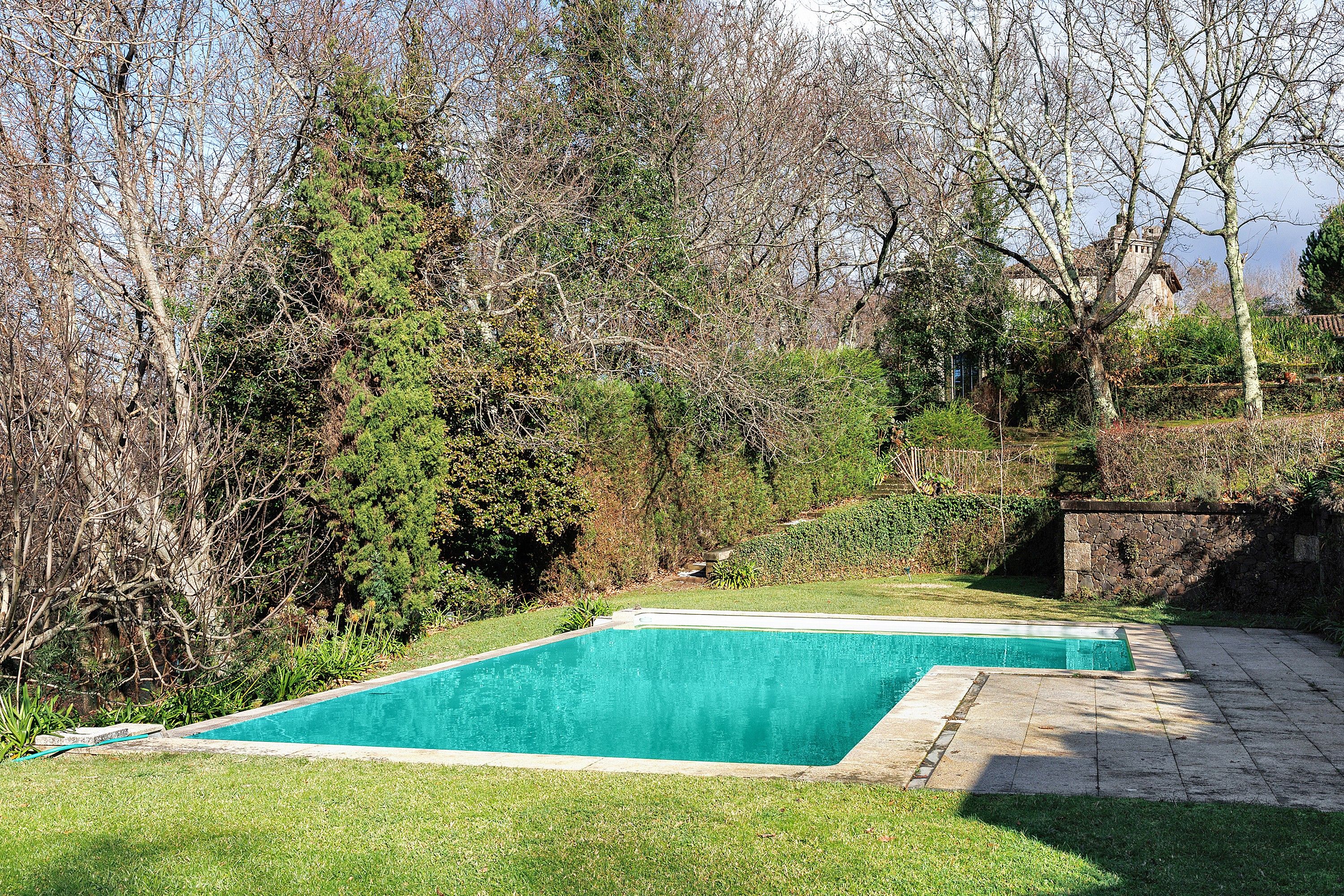Ein rechteckiger Swimmingpool mit einer Steinterrasse, eingebettet in eine Rasenfläche mit Bäumen und einer Steinmauer im Hintergrund.