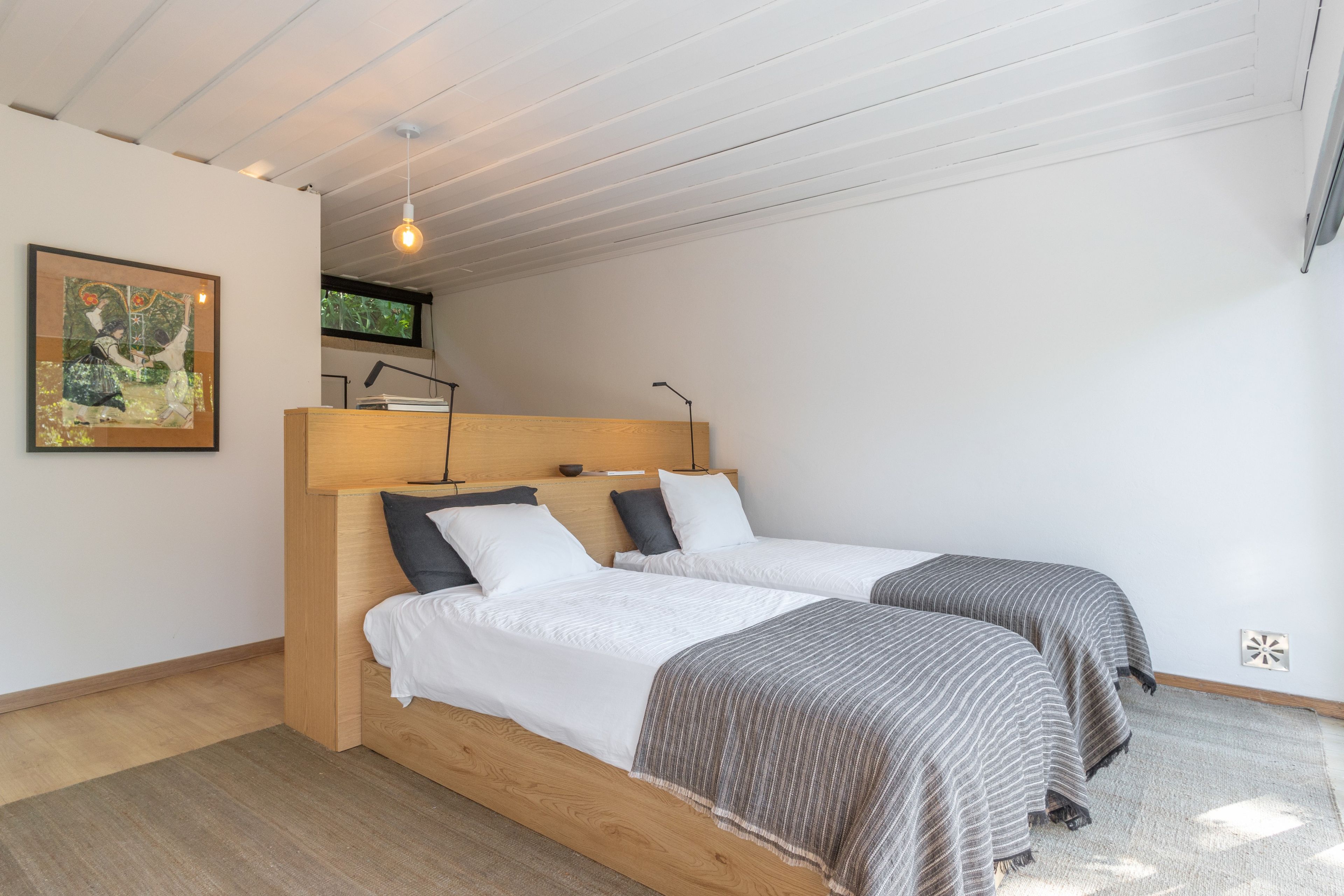 Une chambre avec deux lits, une tête de lit en bois, un tableau encadré au mur et une fenêtre donnant sur un paysage verdoyant.