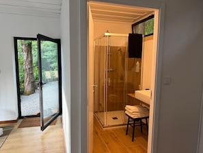 Ein modernes Badezimmer mit Duschkabine aus Glas, einer Holzbank und Blick ins Freie durch eine Glastür.