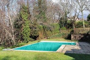 Een rechthoekig zwembad met een omliggend stenen terras, gelegen in een gazon met bomen en een stenen muur op de achtergrond.