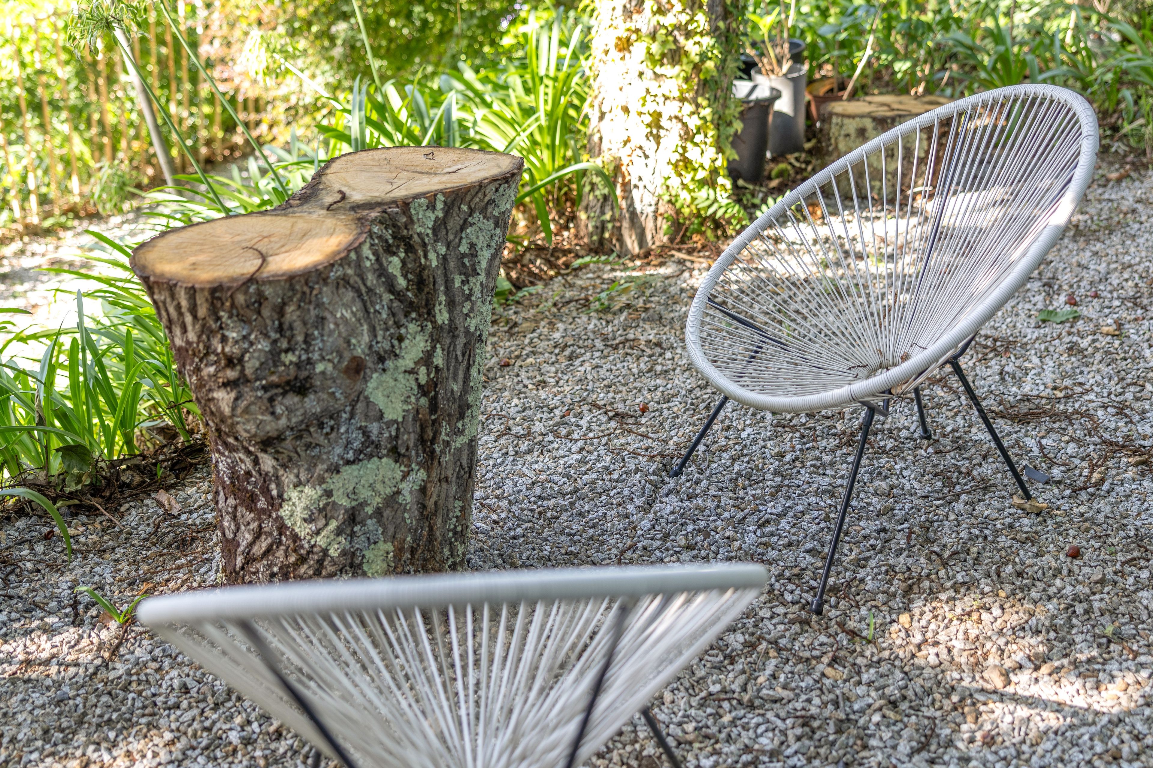 Ein Gartenbereich mit einem weißen Metallstuhl, einem hölzernen Baumstumpf und einem Metalltisch.