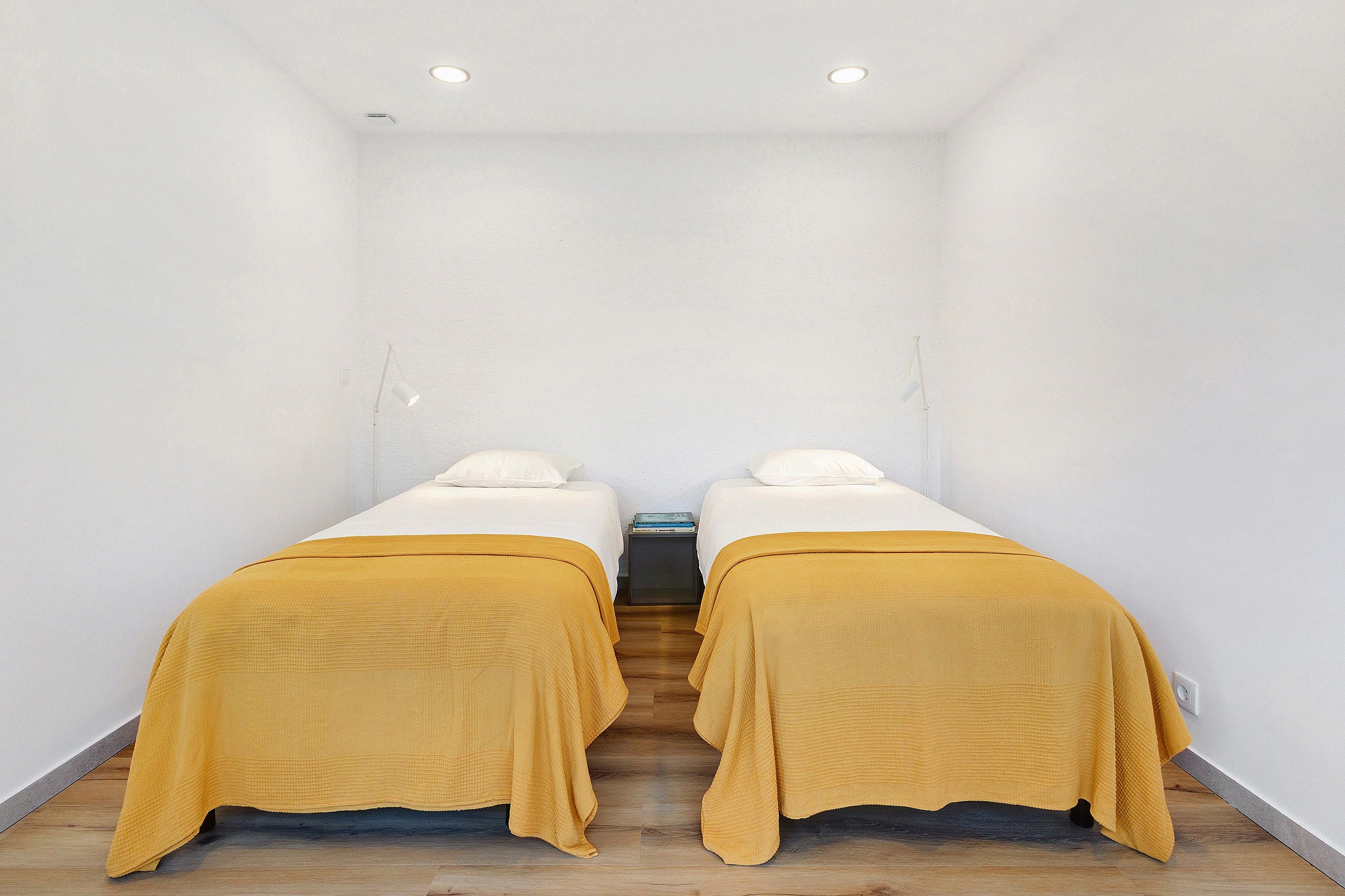 Una habitación con dos camas, cada una con un colchón blanco y una colcha color mostaza, y una mesita de noche pequeña entre ellas.