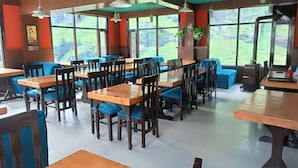 Un restaurante con mesas de madera y sillas con cojines azules, ventanas amplias con vistas a la vegetación y lámparas colgantes.