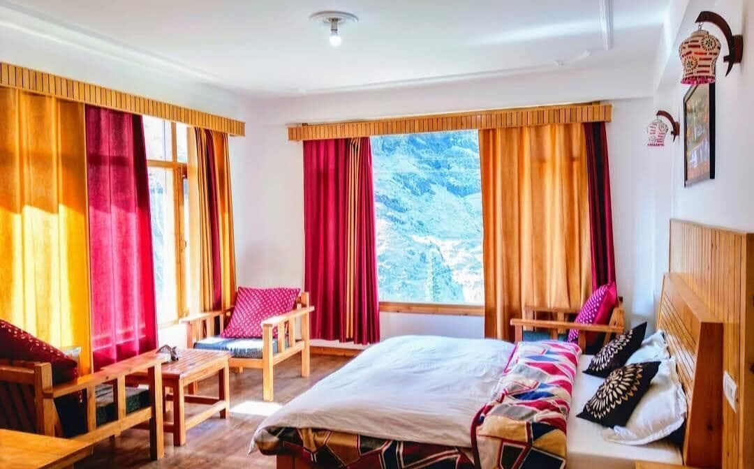 Una habitación con cama, dos sillas, una mesita y ventanas grandes con cortinas.