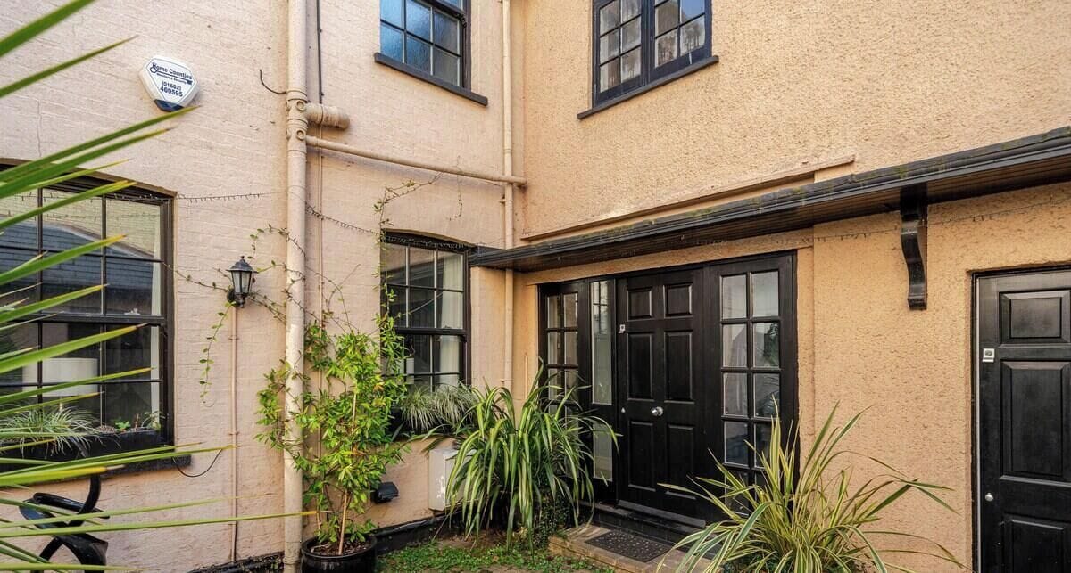 A building with a beige exterior, black-framed windows, and a black door. There are potted plants and a small garden area.
