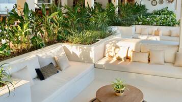 A modern outdoor living area with white sofas and a round wooden table, surrounded by lush greenery and decorative wall art.