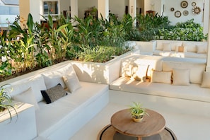 Un espacio exterior moderno con sofás blancos y una mesa redonda de madera, rodeado de abundante vegetación y detalles decorativos en las paredes.