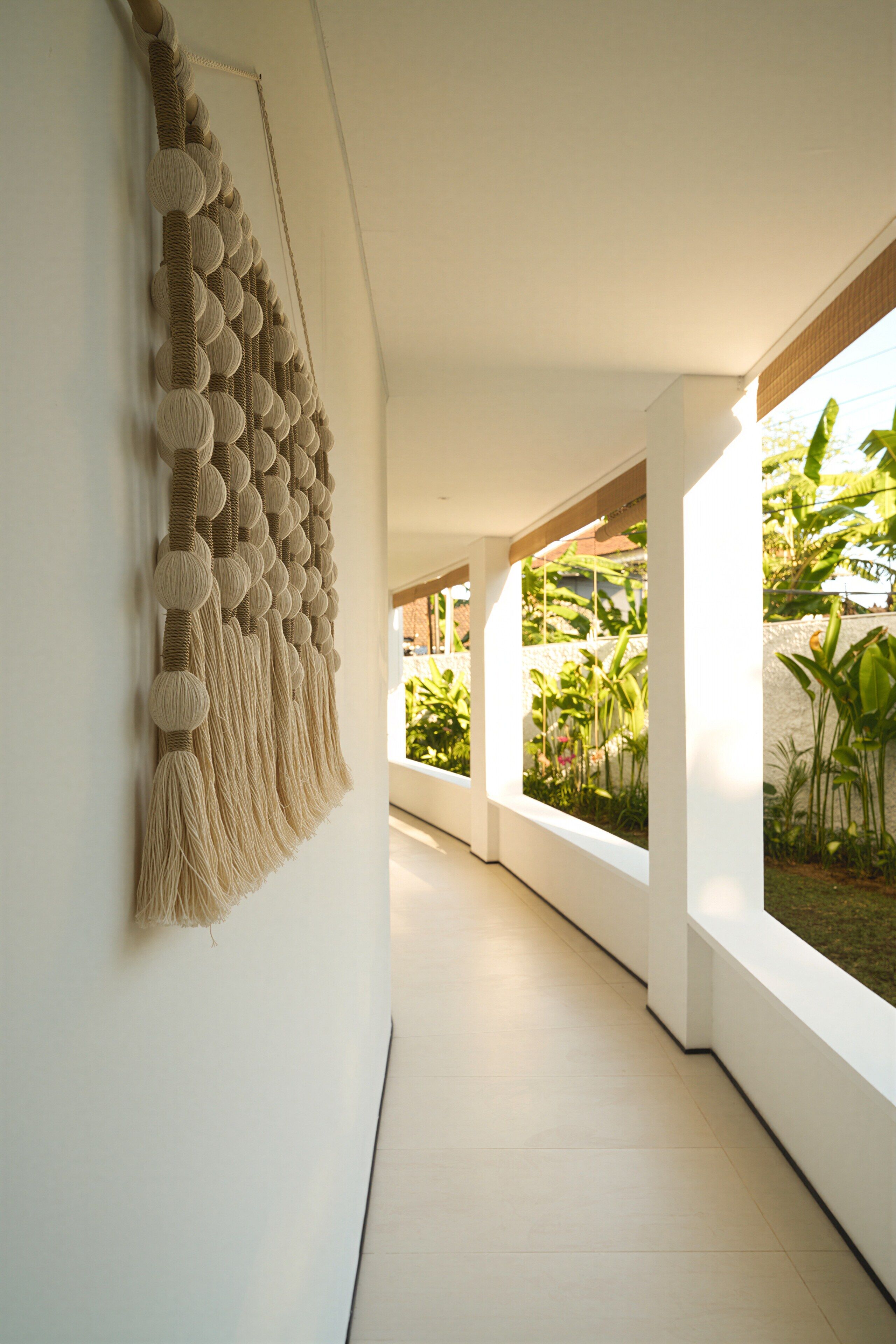 A corridor with a macramé wall hanging, white walls, and large windows with greenery outside.