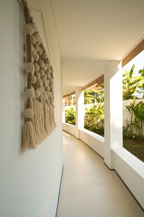 Un pasillo con un tapiz de macramé en la pared, paredes blancas y ventanas amplias con follaje en el exterior.