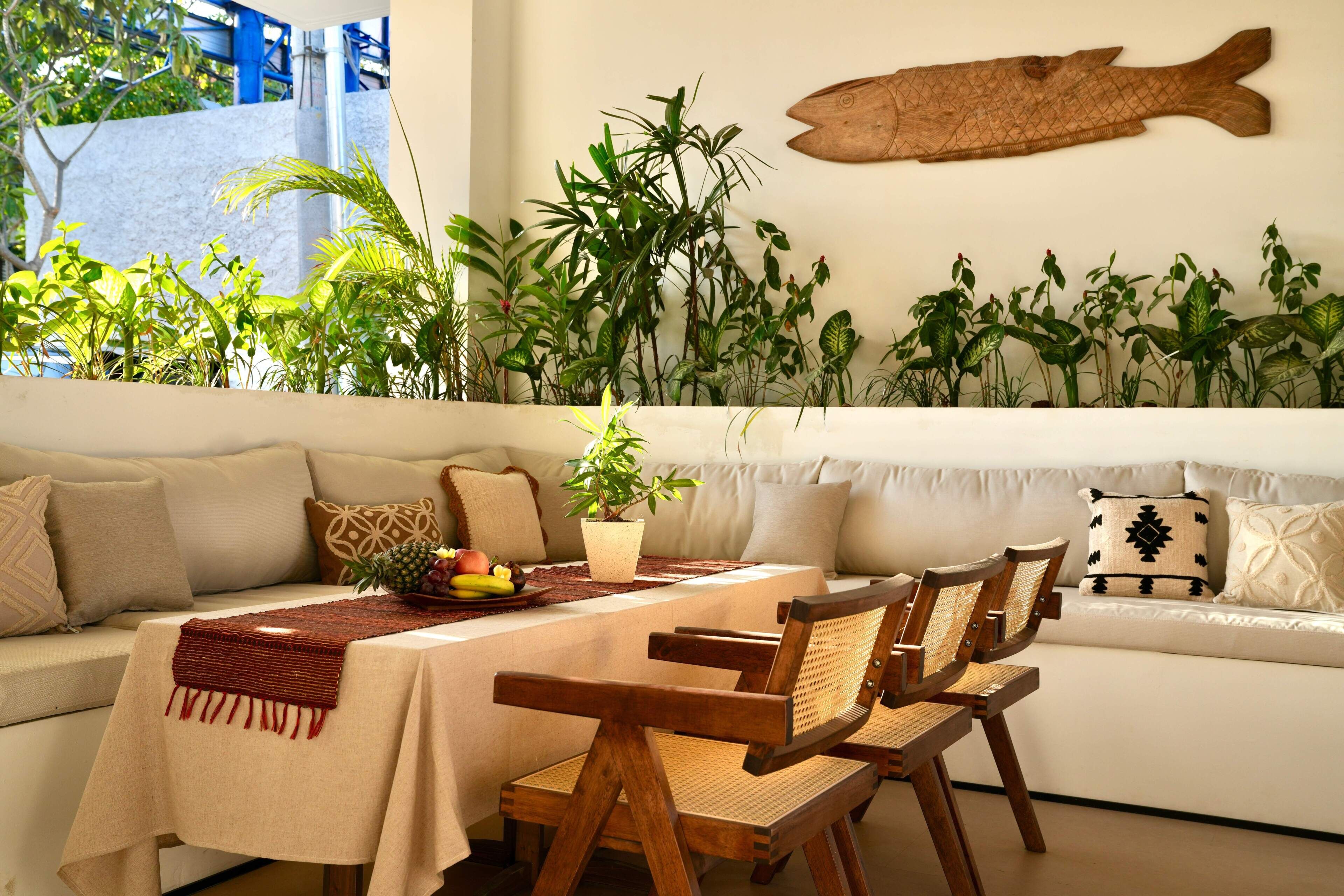 A dining area with a table set for two, surrounded by wooden chairs with woven seats. A fruit bowl is on the table, and there is a potted plant on the floor.