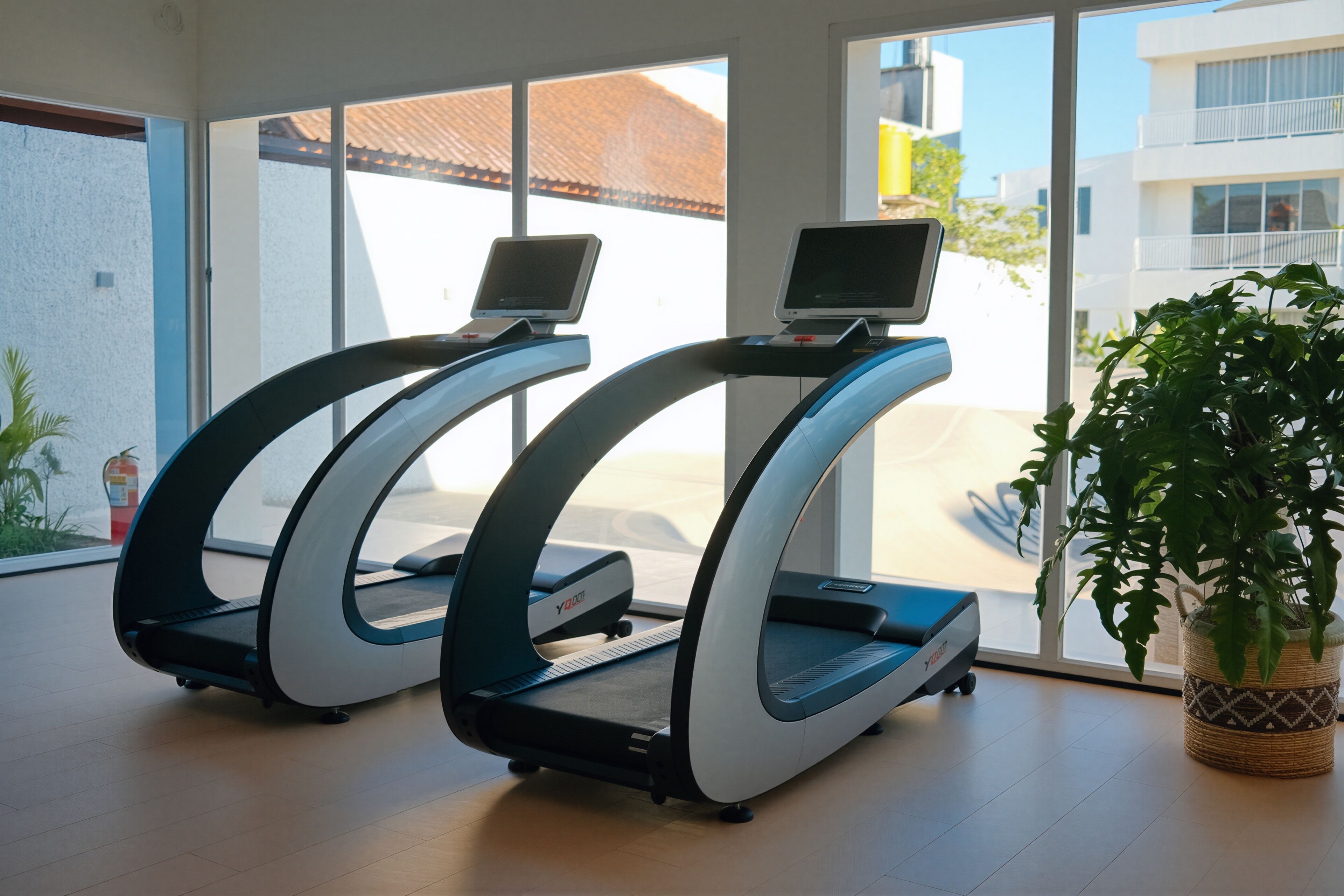 Deux tapis de course équipés d’écrans numériques, placés dans une pièce dotée de grandes fenêtres et agrémentée d’une plante en pot.