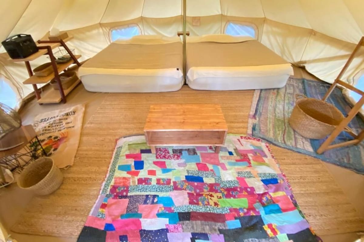A tent interior with a quilted floor mat, a wooden bench, and a woven basket.