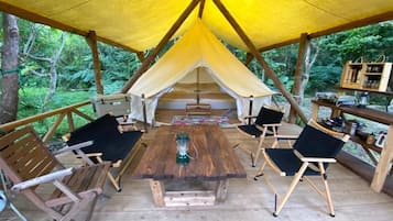 Een houten terras met een centrale tafel en stoelen, omringd door een gele tentconstructie.