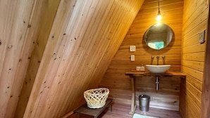 Interior de madeira com teto inclinado, um espelho redondo, uma pia e um pequeno banquinho.