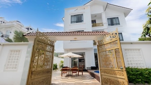 A gated entrance with ornate golden gates, a checkered outdoor seating area, and a white building with balconies in the background. - Luxury An Home Beach Villa (Rach Dua)
