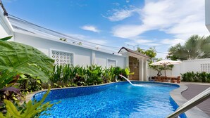 A swimming pool with a tiled edge, surrounded by lush greenery and a white building with decorative windows. - Luxury An Home Beach Villa (Rach Dua)
