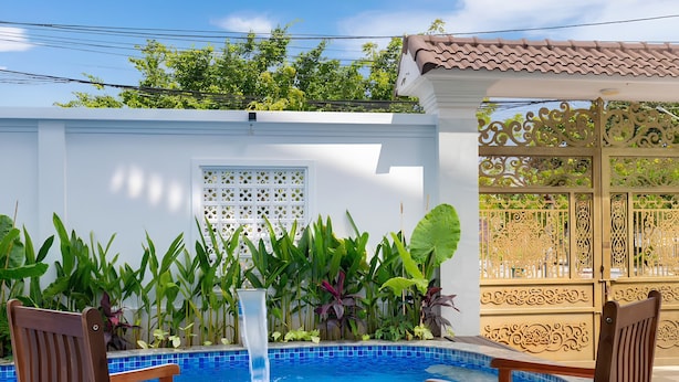 A poolside area with two wooden chairs and a table, surrounded by lush greenery and a white wall. - Luxury An Home Beach Villa (Rach Dua)