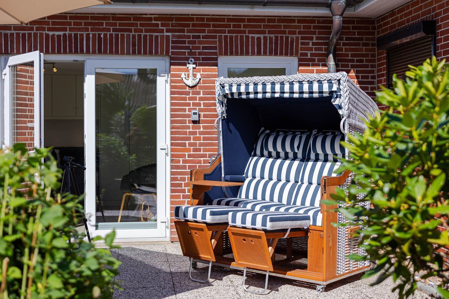 A sunlit outdoor seating area with a wooden bench and striped cushions, a brick wall, and a sliding glass door.