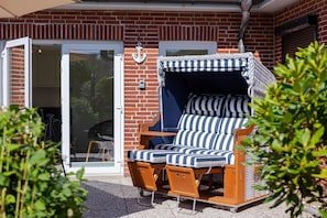 Ein sonniger Außenbereich mit Sitzgelegenheit, einer Holzbank und gestreiften Kissen, einer Ziegelwand und einer Schiebetür aus Glas.
